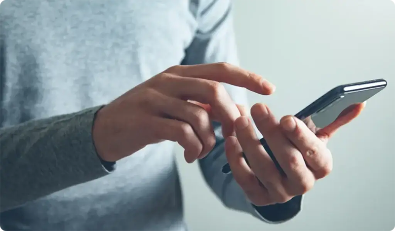 A person wearing a grey long-sleeve shirt uses a smartphone, holding it in one hand and tapping the screen with the other—perhaps engaging with resources from Troomi Parent Academy. The background is blurred and neutral.