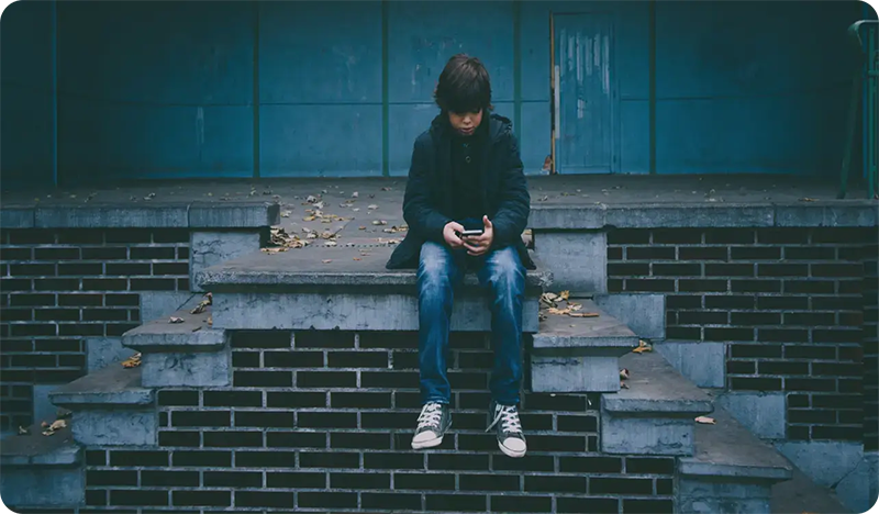 A boy in a dark jacket and jeans sits alone on concrete steps, looking down at his phone—a scene that echoes the importance of digital safety discussed in troomi parent academy—surrounded by scattered leaves and a blue wall in the background.