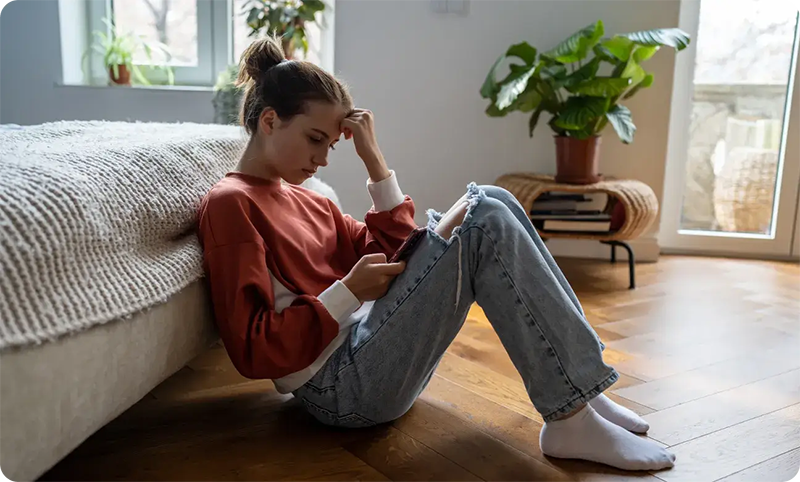 A young woman sits on a wooden floor by a bed, looking at her phone with a thoughtful expression. She wears a red sweater, ripped jeans, and white socks. A potted plant and books are nearby by a bright window, suggesting she might be exploring resources like the Troomi Parent Academy to learn more about healthy technology habits.