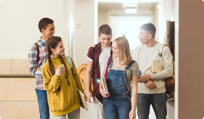 Five smiling teenagers walk together in a hallway, holding books and backpacks, appearing to be students engaged in friendly conversation—perhaps discussing what they learned in troomi parent academy.