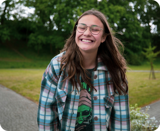 A young person with long brown hair and glasses smiles widely, wearing a plaid jacket and standing outdoors on a cloudy day with trees and grass in the background.