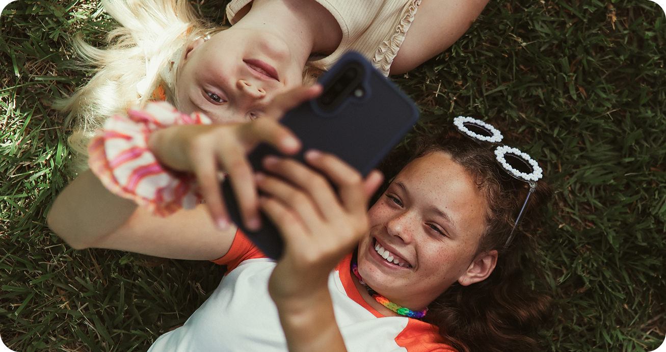 Two girls lying on the grass, smiling and taking a selfie with a smartphone. One wears sunglasses on her head and a colorful necklace, while the other has a scrunchie on her wrist.