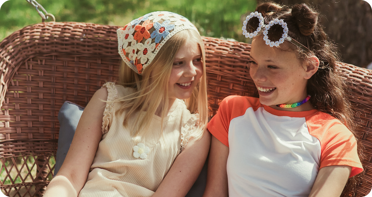 Two girls sit on a wicker bench outdoors, smiling at each other. One wears a floral headscarf and beige dress; the other has flower-shaped sunglasses, a colorful choker, and an orange and white shirt. Green grass is visible in the background.