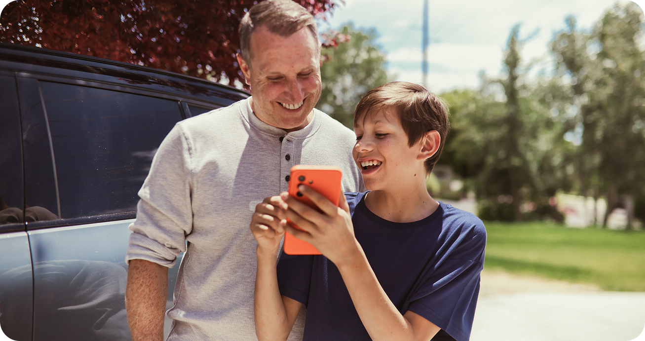 A smiling adult and child stand outdoors by a car, laughing together while looking at a bright orange smartphone. Sunlight filters through trees in the background.