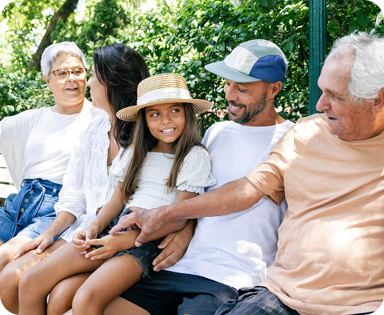 Five people of varying ages sit closely on a bench outdoors, smiling and talking. A girl in a straw hat sits on a mans lap, surrounded by adults. Green foliage fills the background.