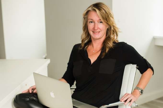 Larissa Mills is shown wearing a black top, and sitting in a white chair with a silver MacBook on her lap.