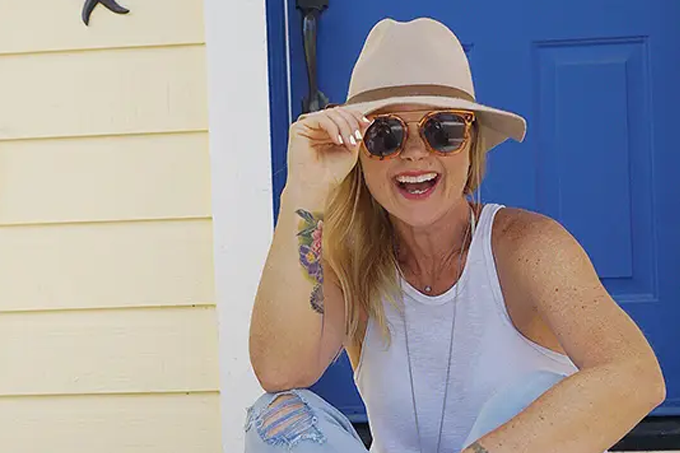 Fresh Start Family's Wendy Snyder is shown wearing a hat and sunglasses as she sits on steps by a bright blue door and yellow wall, smiling and holding her sunglasses. She has a floral tattoo on her arm and wears a white tank top and ripped jeans.
