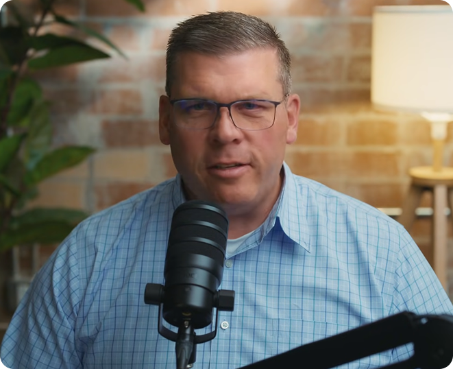 A man in a light blue checkered shirt speaks into a studio microphone.