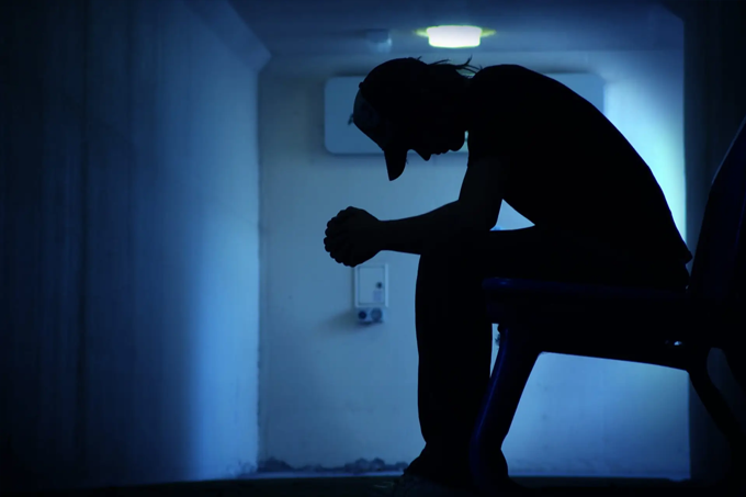 A person sits on a chair in a dimly lit hallway, hunched forward with elbows on knees and hands clasped, appearing deep in thought or sad.