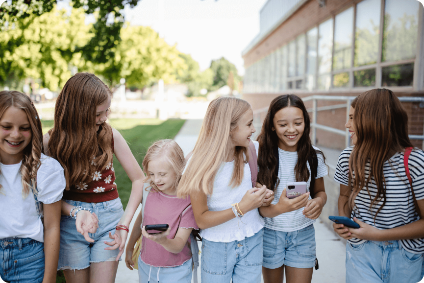 A group of six young girls stand on a sidewalk outside a building, laughing and looking at their Troomi smartphones. They are dressed in casual summer clothes, and the background features trees alongside a glass-windowed building.